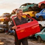 Mécanicien de 55 ans dans une casse automobile à Aix les Milles, tenant une pièce de voiture rouge vive parmi des piles de pièces colorées