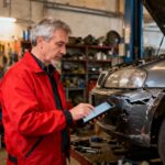 Expert automobile en veste rouge examinant les dommages d'une voiture avec une tablette de diagnostic dans un atelier