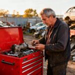Homme de 50 ans examinant des pièces de voiture dans une casse automobile à Orléans, avec une boîte à outils rouge