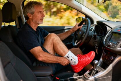 Homme d'âge mûr conduisant une voiture avec une chaussure rouge visible sur son pied opéré, éclairage naturel de l'habitacle