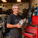 Mécanicien de 55 ans tenant une pièce de voiture recyclée devant une boîte à outils rouge vif