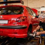 Peugeot 206 rouge avec modifications sportives dans un atelier, homme ajustant l'échappement sous éclairage chaud