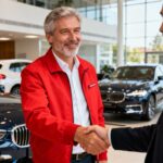 Vendeur automobile souriant vêtu d'une veste rouge serre la main d'un client dans un showroom de concession auto